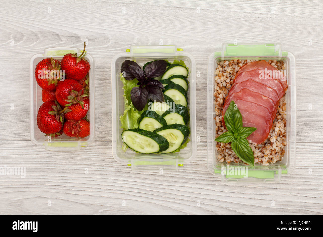 Kunststoff Mahlzeit prep Container mit frischen Erdbeeren, Gurken und Salat, gekochten Buchweizen Brei und Scheiben von Fleisch auf grau Holz- Hintergrund. Top Stockfoto