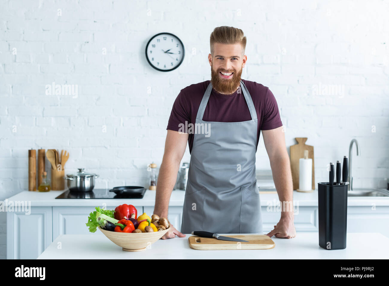 Gut aussehender bärtiger junger Mann in Schürze lächelte Kamera beim Kochen in der Küche Stockfoto