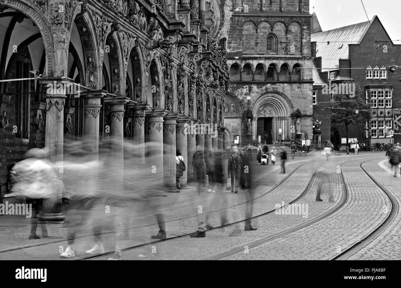Bremen, Deutschland - Verschwommenes menschliche Figuren die Straße und Straßenbahnschienen überqueren vor dem historischen Rathaus und der Kathedrale (lange Belichtung, monochrom Stockfoto
