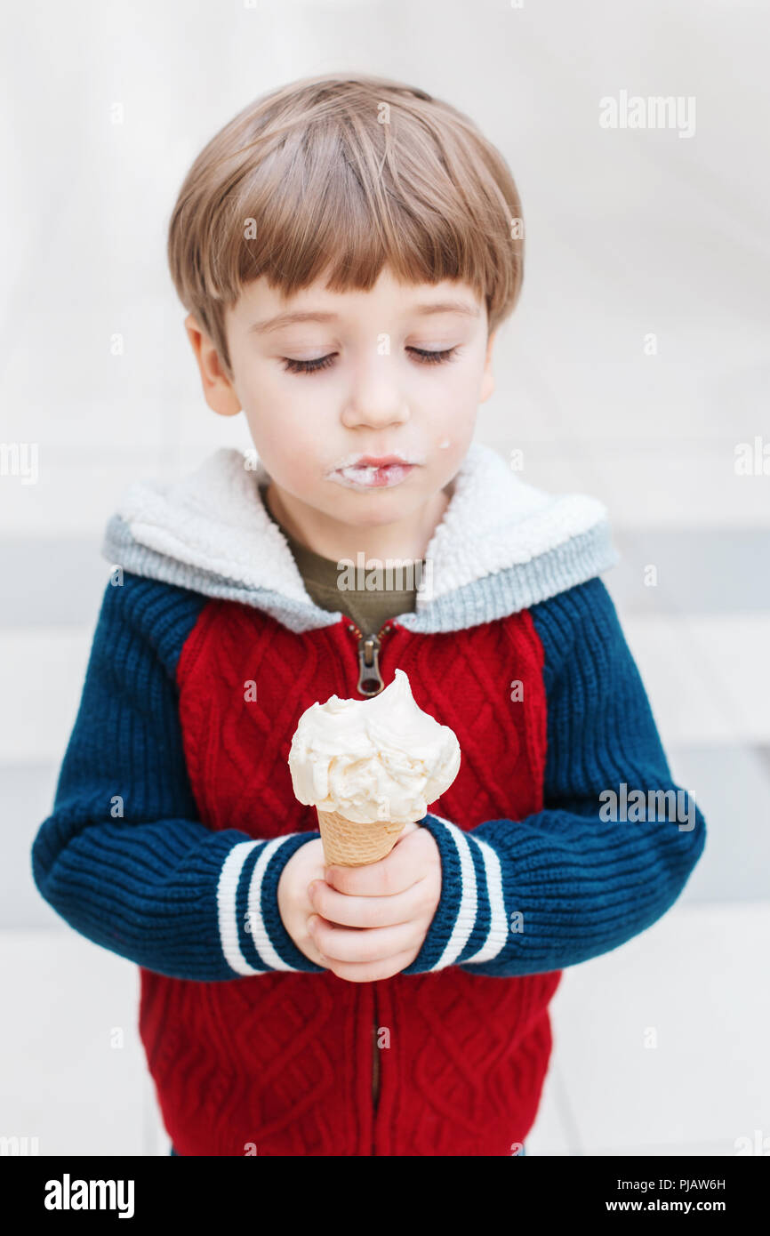 Closeup Portrait von niedlichen weißen Kaukasischen blond Vorschule Junge mit grünen Augen in rot pullover Essen lecken Eis in der Waffel Kegel. Stockfoto