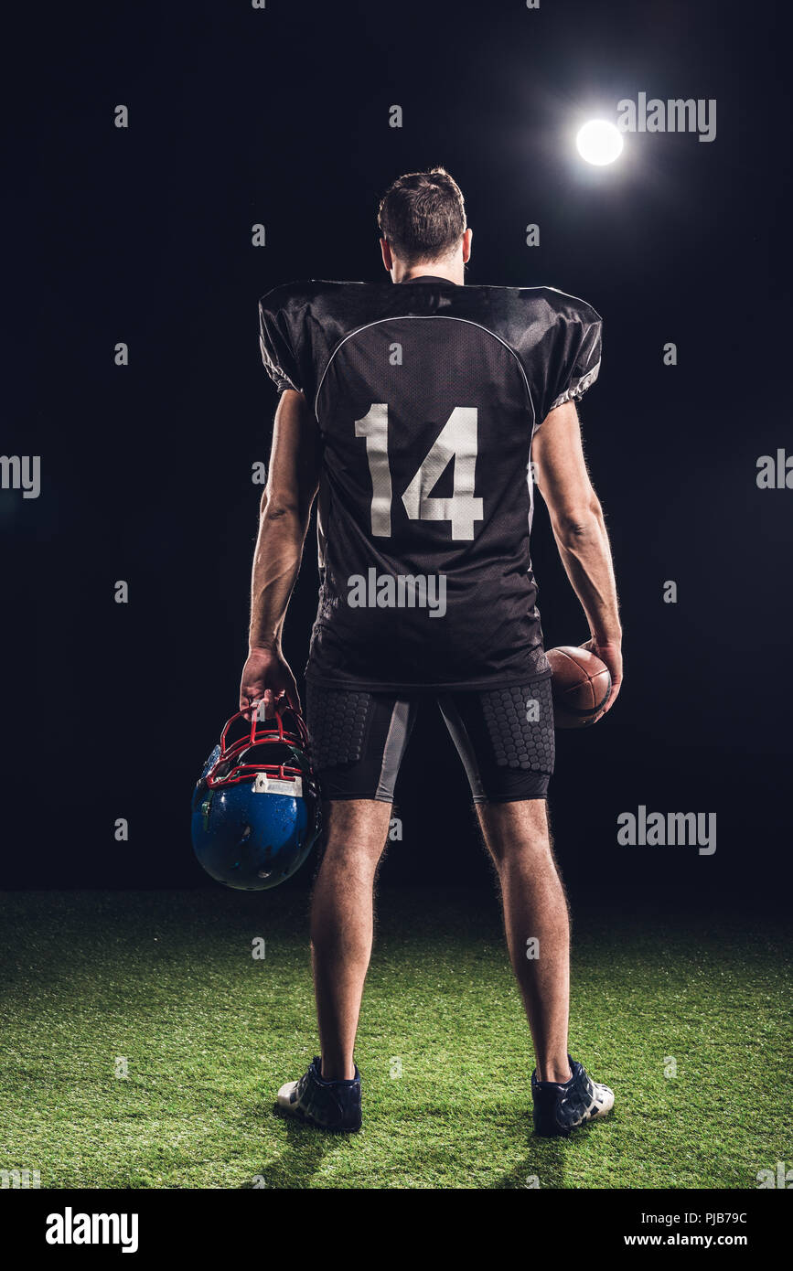 Ansicht der Rückseite des American Football Spieler mit Ball und Helm stehen auf Gras unter Spotlight auf Schwarz Stockfoto