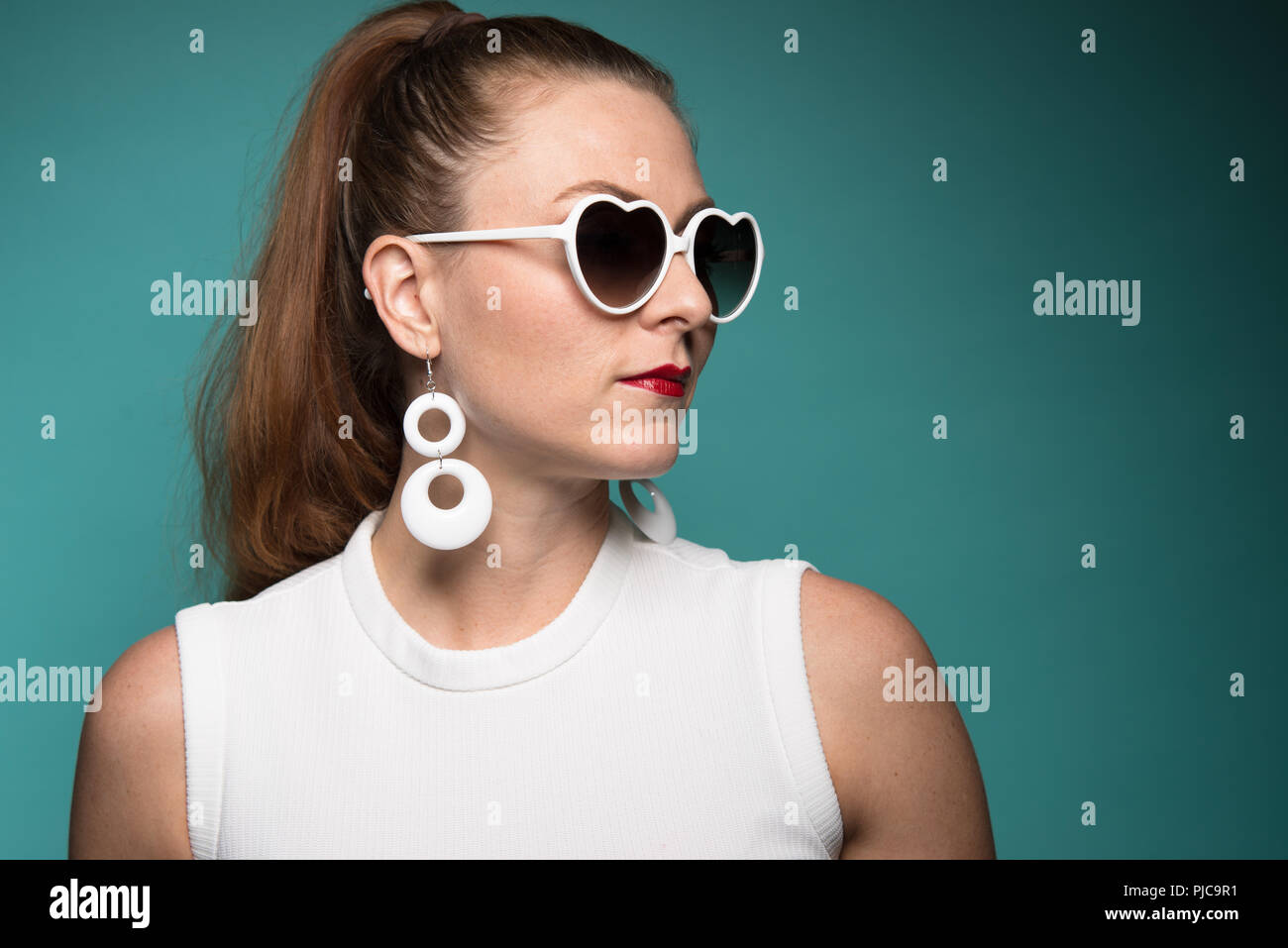 Ein rotes Haar kaukasische Frau mit Pferdeschwanz, trug einen Mod 60s style Ohrringe und herzförmige sungalsses posiert, eine Nahaufnahme Porträt. Stockfoto
