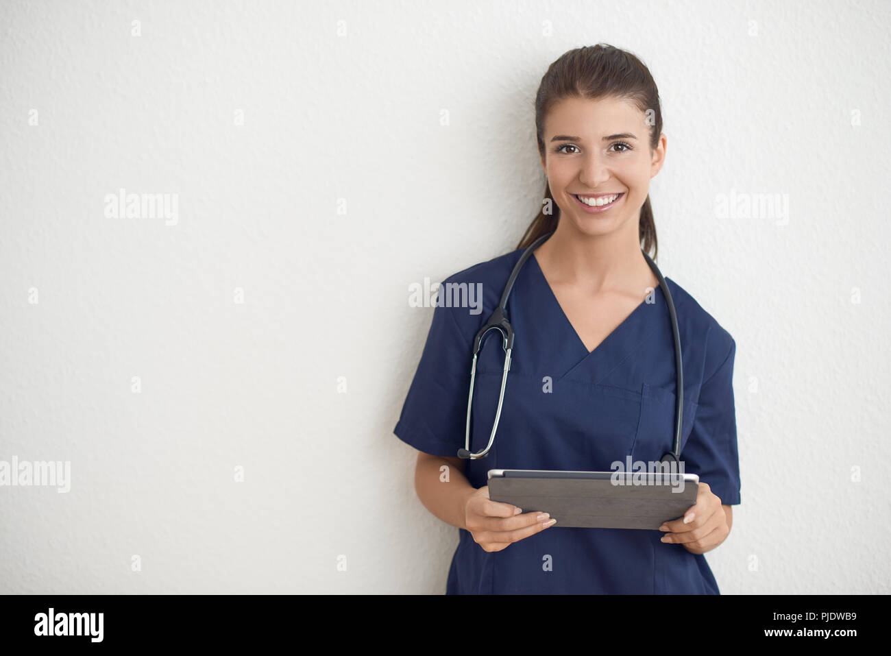 Junge Frau Arzt oder Krankenschwester in Blau scrubs Holding ein Tablet PC vor einer weißen Wand Stockfoto