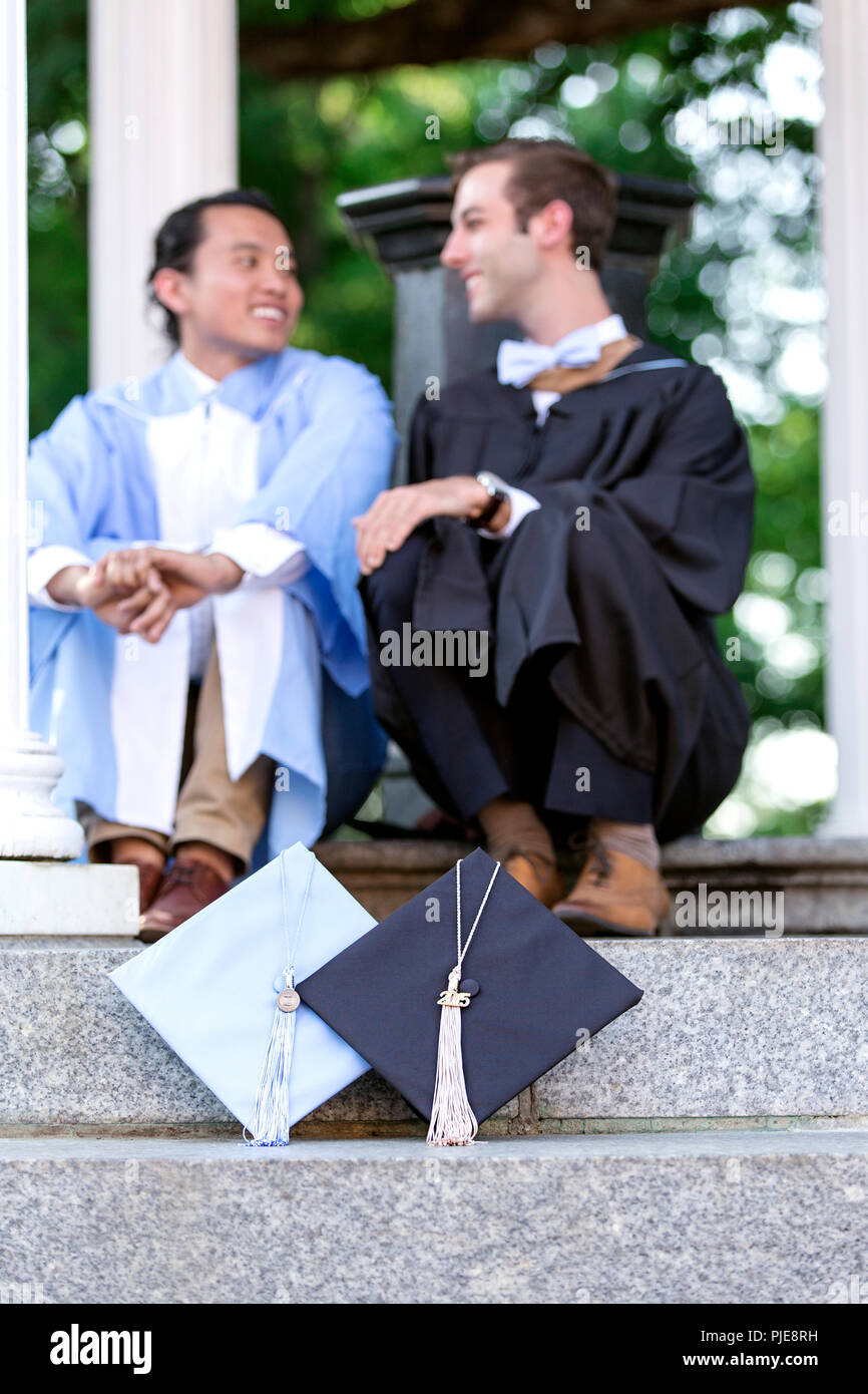 Zwei männliche Hochschulabsolventen sitzen auf der Treppe im Gespräch mit mortarboards im Vordergrund positioniert Stockfoto