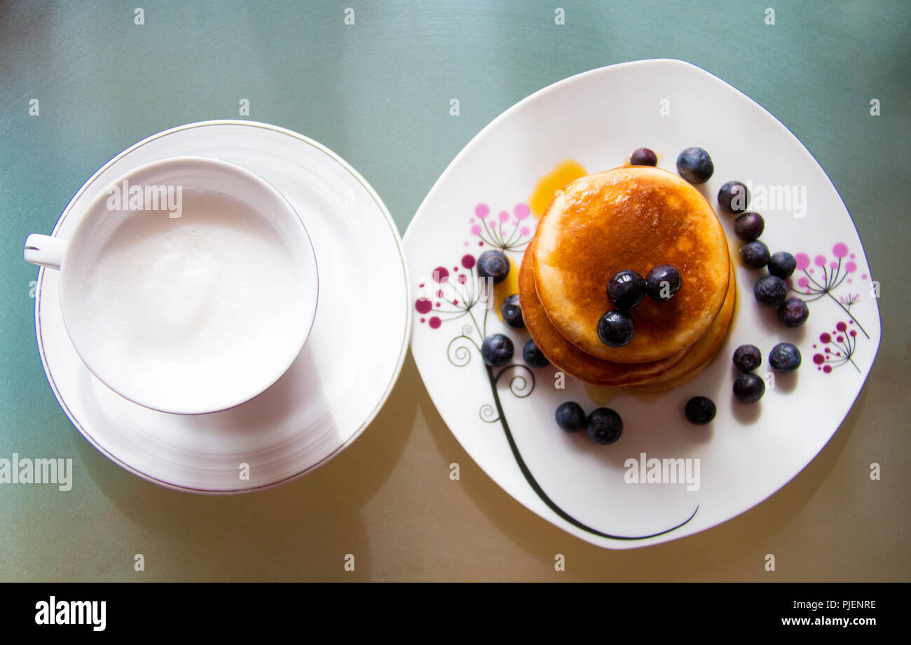 Oben auf der Platte mit Tasse cremigen Kaffee und die Platte mit der Süße Pfannkuchen mit Heidelbeeren Früchte und Ahornsirup, geschmackvollen Frühstück MAHLZEIT Stockfoto