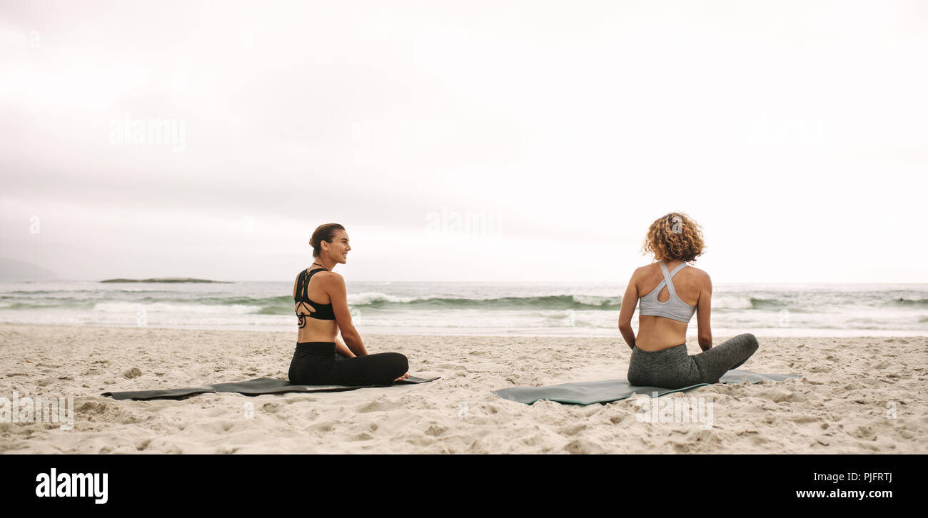 Rückansicht zweier Frauen in Fitnesskleidung, die Yoga üben, am Strand sitzen. Fitness-Frauen sitzen auf Yogamatten und machen Yoga mit Blick auf das Meer. Stockfoto