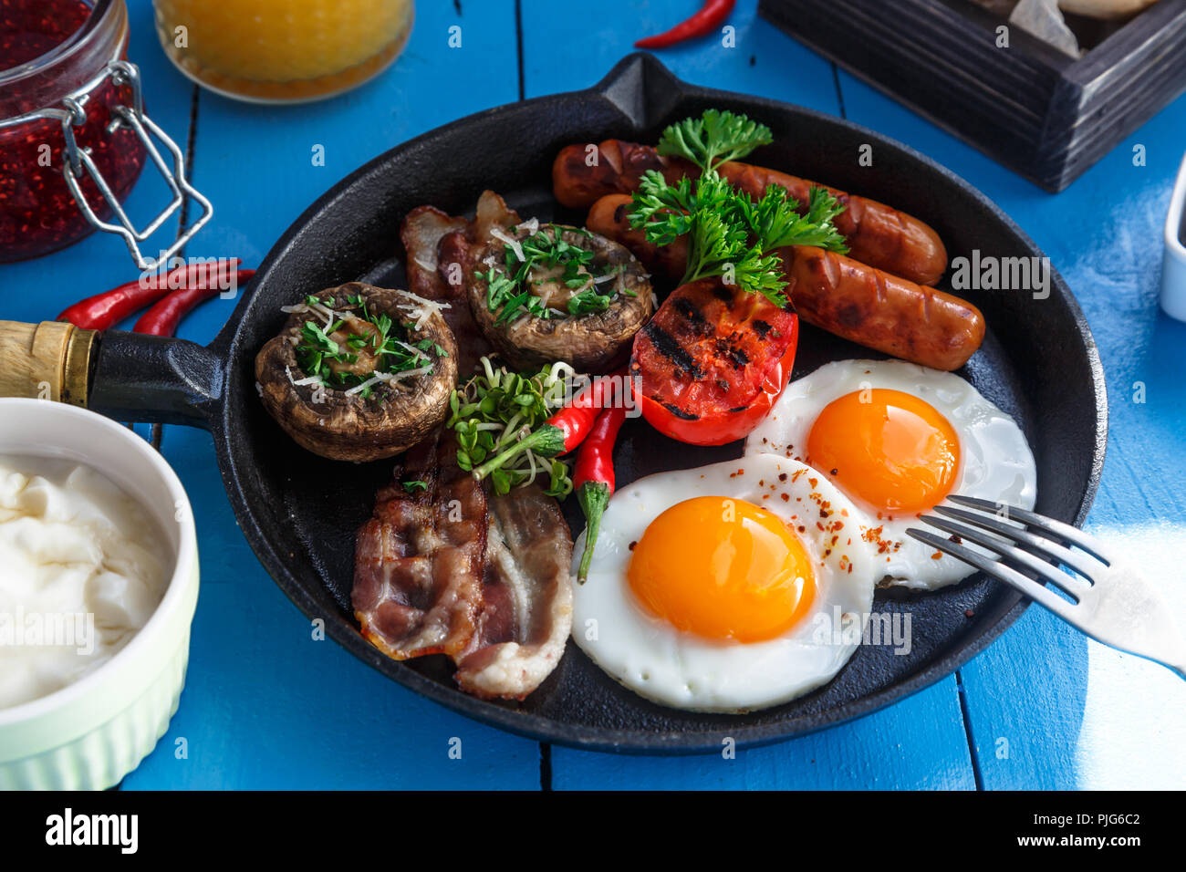 Ansicht schließen Englisches Frühstück in einer Pfanne mit Spiegelei, Würstchen, Speck, Pilzen, Marmelade und Orangensaft auf hölzernen Hintergrund Stockfoto