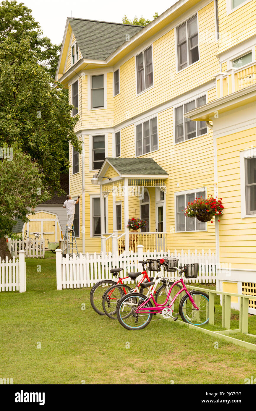 Die Außenseite des charmanten Gelb home auf dem historischen Mackinac Island. Mann auf Leiter Malerei Haus. Fahrräder geparkt sind das wichtigste Transportmittel. Stockfoto