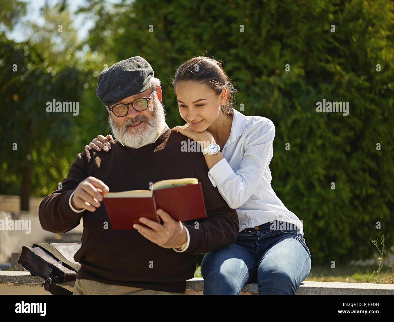 Helfen Sie jemandem, den Sie lieben. Portrait der junge lächelnde Mädchen umarmt Großvater mit Buch gegen City Park. Gemeinschaft und Familie lifestyle Konzept Stockfoto