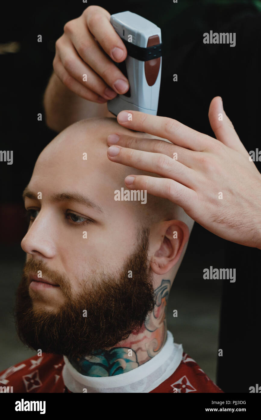 Ein gut aussehender Bärtiger skinhead Mann in einem Friseursalon. Der barbier rasiert seinen Kopf mit einem elektrischen Trimmer. Stockfoto