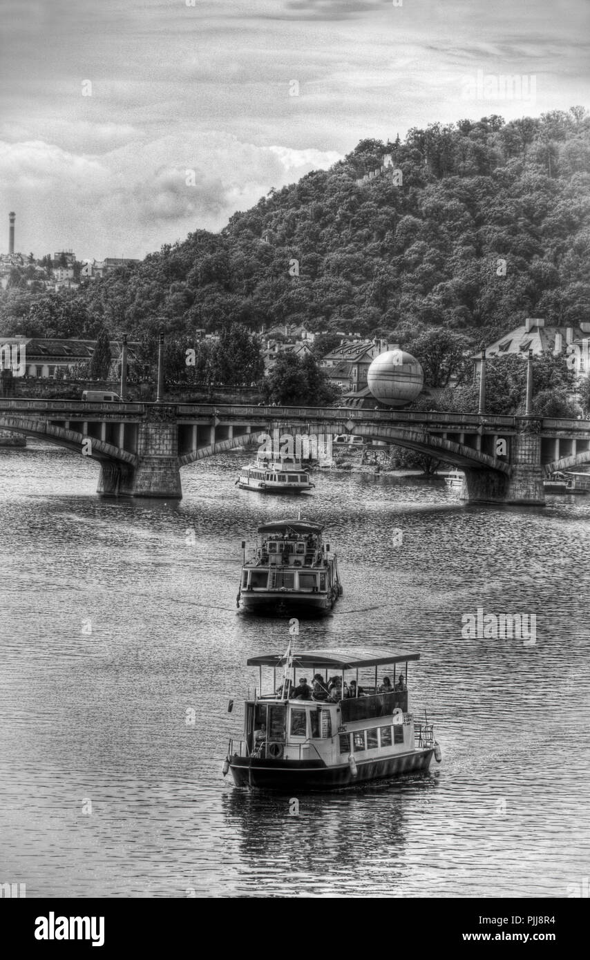 Blick über den Fluss Moldau, Prag mit drei Schiffen und eine Brücke und Wald im Hintergrund, Foto in Schwarz und Weiß, dramatische Stockfoto