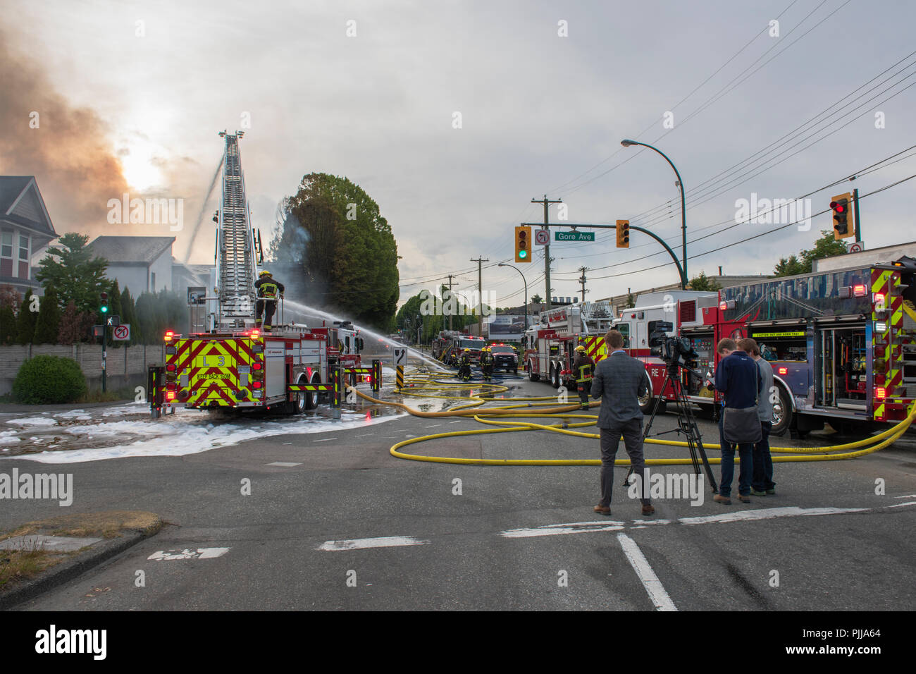 Reporter im Vordergrund der Berichterstattung über die Löschfahrzeuge und feuerwehrleute an der Szene eines Hauses Feuer, Vancouver city. Stockfoto