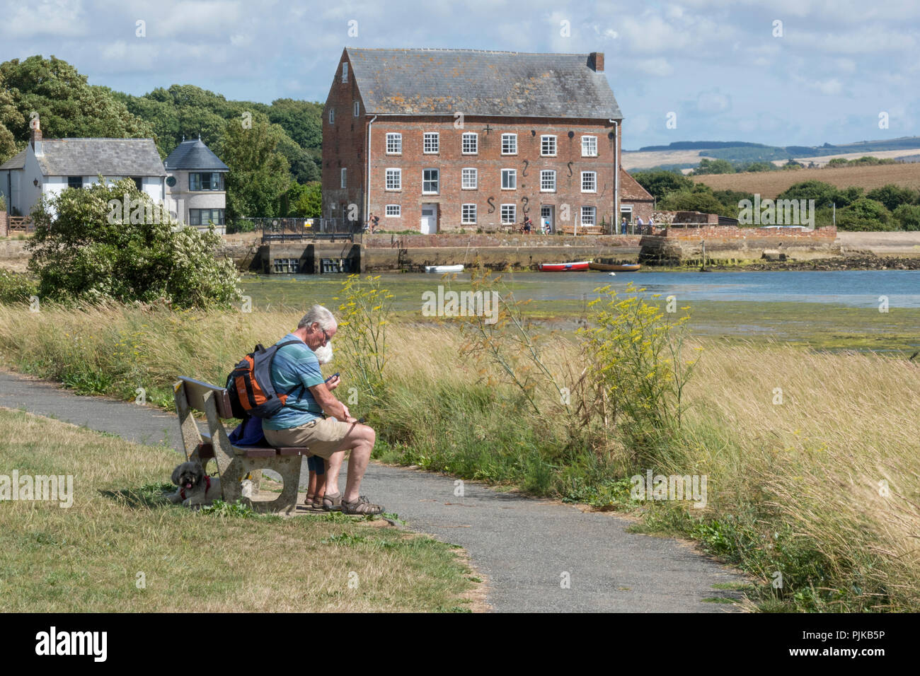 Walker oder Wanderer sitzen auf einer Bank an der Seite des Fluss Yar in Yarmouth auf der Isle of Wight mit der Tide Mill in der Ferne oder Hintergrund. Stockfoto