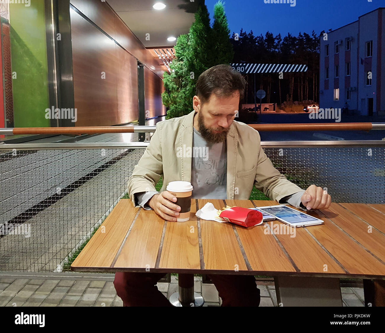 Ein bärtiger Mann mittleren Alters der kaukasischen Aussehens sitzt an einem Tisch in einem Sommer Cafe am Abend Aufpassen der Nachrichten auf dem Bildschirm eines Tablet, trinken Kaffee und essen Pommes frites. Stockfoto