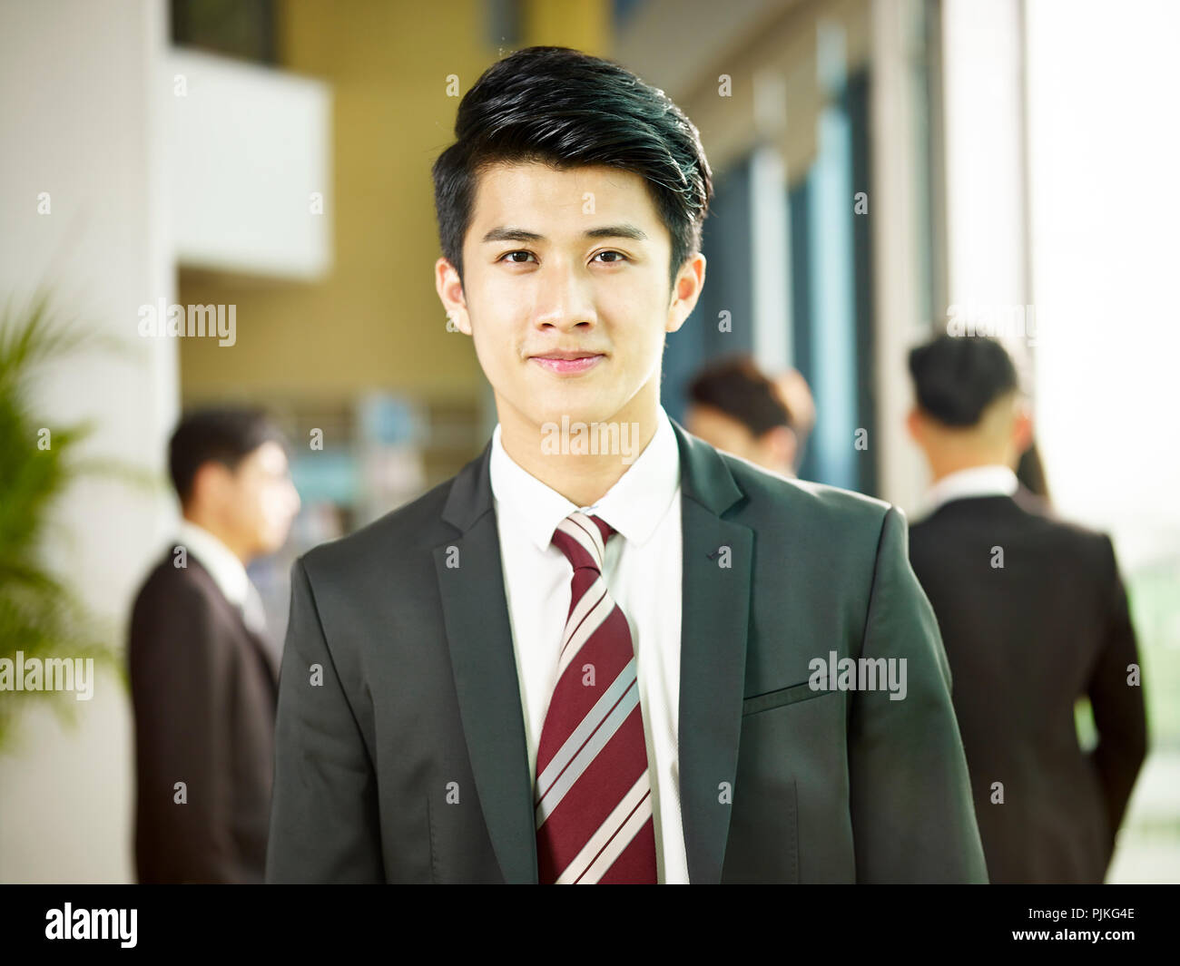 Porträt einer jungen asiatischen Geschäftsmann steht in der Lobby des modernen Bürogebäude an der Kamera schaut lächelnd. Stockfoto