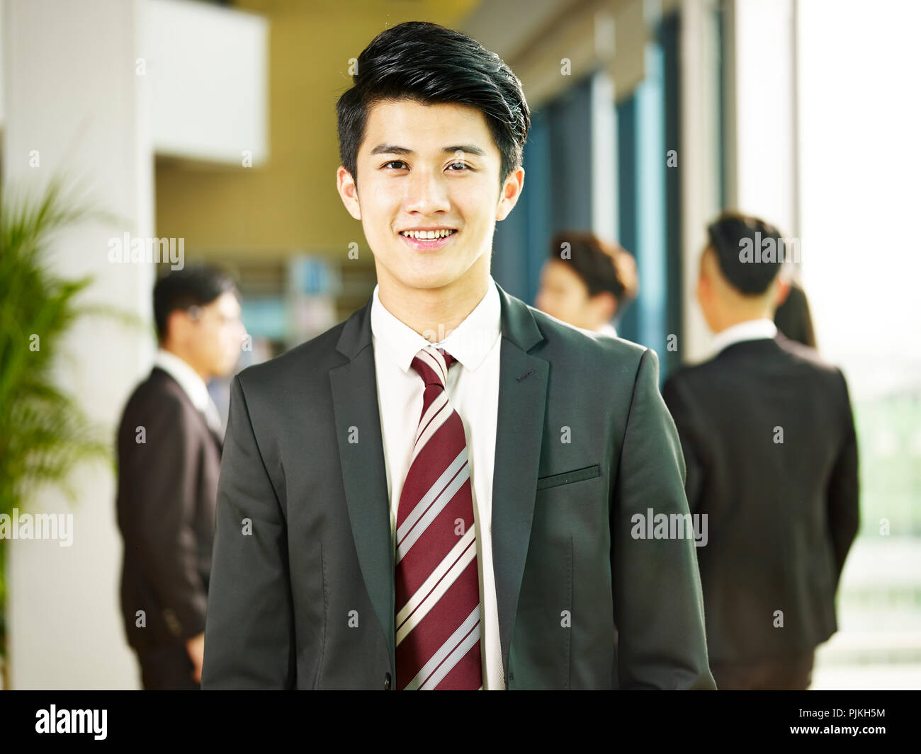 Porträt einer jungen asiatischen Geschäftsmann steht in der Lobby des modernen Bürogebäude an der Kamera schaut lächelnd. Stockfoto