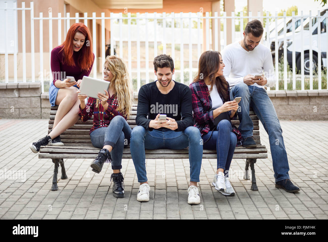Gruppe von Jugendlichen mit Smartphones und Tablet Pcs im Freien im städtischen Hintergrund. Frauen und Männer sitzen auf einer Bank in der Straße tragen casua Stockfoto