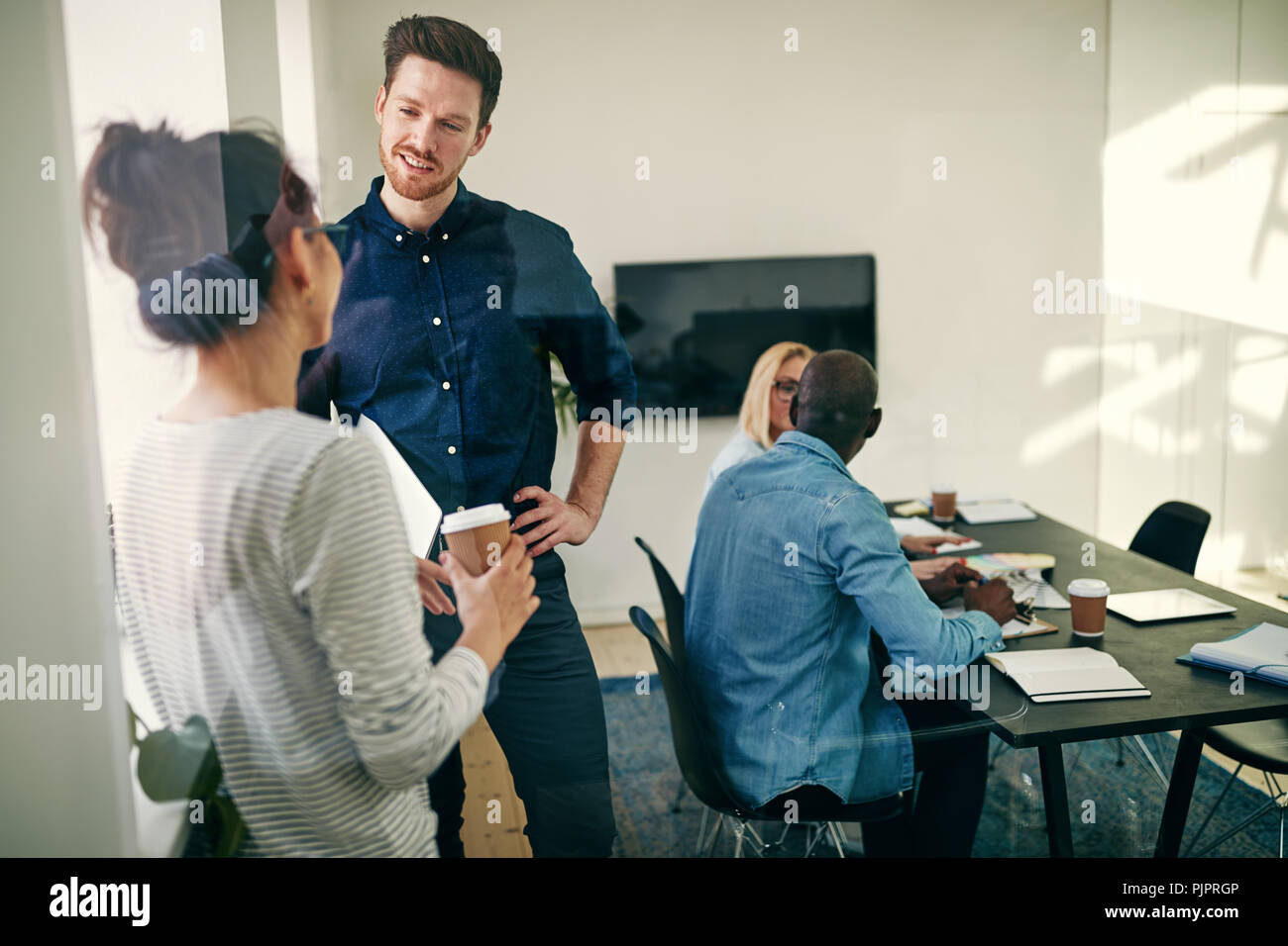 Zwei lächelnde junge Geschäftsleute zusammen gesprochen in einem Büro Boardroom mit Kollegen an einem Tisch im Hintergrund sitzen Stockfoto