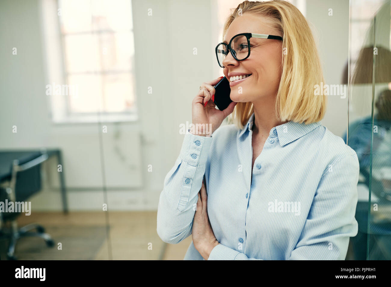 Lächelnden jungen Geschäftsfrau, die in einem modernen Büro im Gespräch über Ihr Handy mit Kollegen in einem Büro hinter ihr zu arbeiten Stockfoto