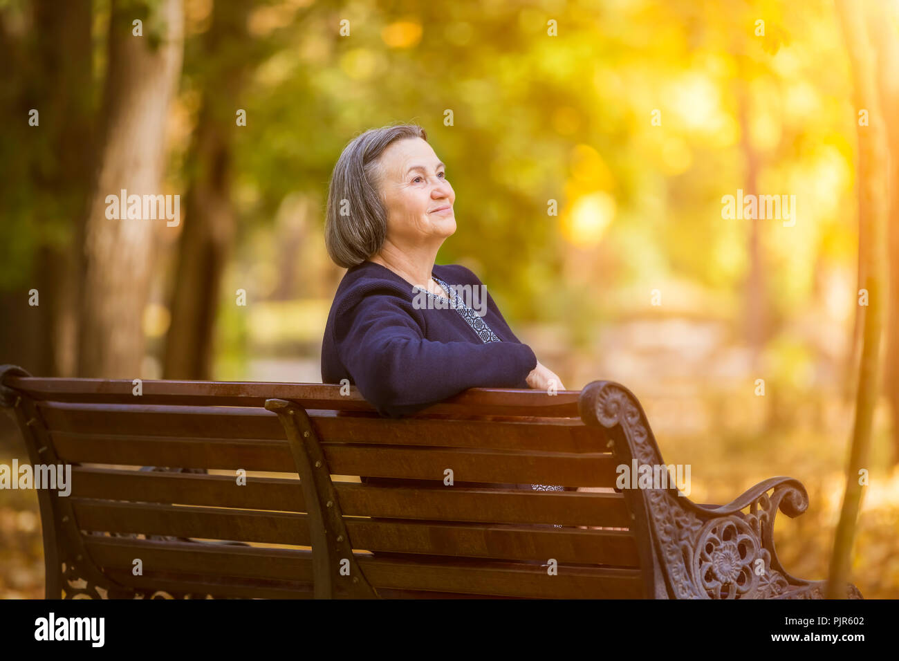 Ältere Frau sitzen auf einer Bank im Herbst Park. Gealterte Frau sitzt auf der Bank im Herbst Park aussieht und lächelnd an der Sonne. Stockfoto