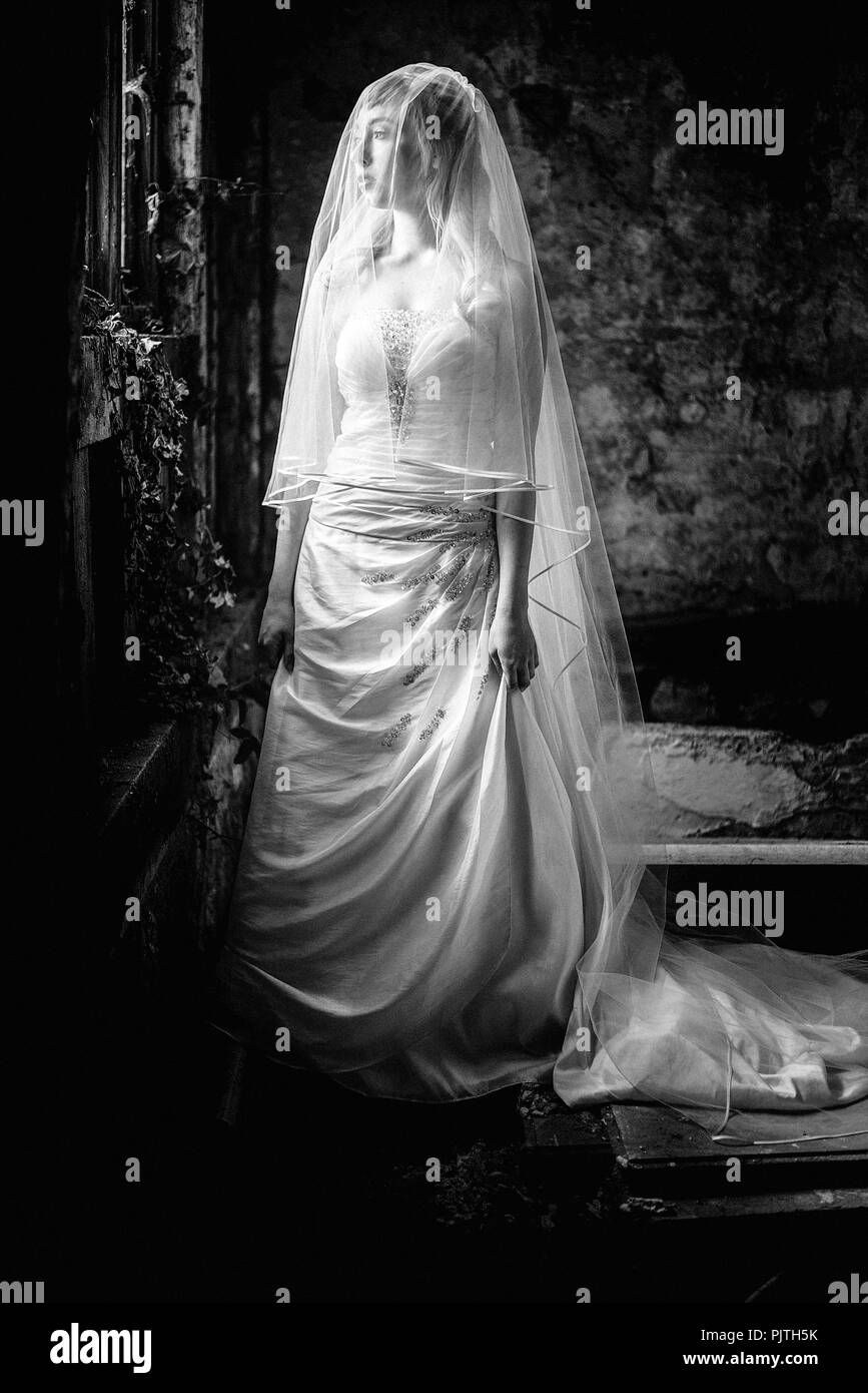 Eine Braut steht in ihrem Hochzeit Kleid und Schleier, schauen aus dem Fenster in einem verlassenen Haus Stockfoto