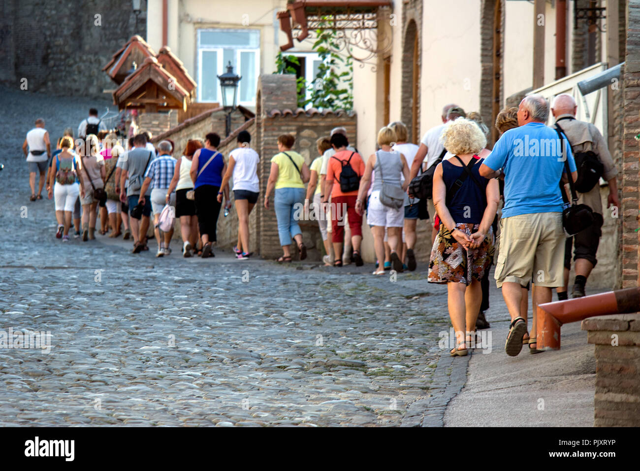 Eine Linie der älteren Touristen aus einem Bus Busreise ordentlich bergauf entlang eines gepflasterten Straße einer Georgischen touristische Stadt. Stockfoto