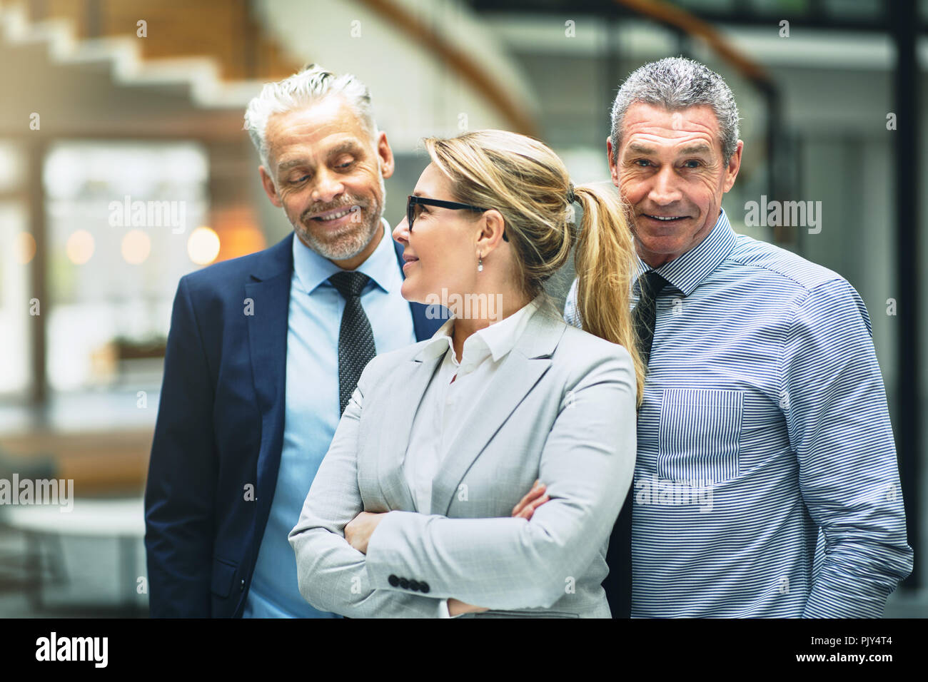 Lächelnd reife Geschäftsfrau im Gespräch mit zwei männlichen Kollegen beim zusammen in der Lobby von einem modernen Bürogebäude stehend Stockfoto