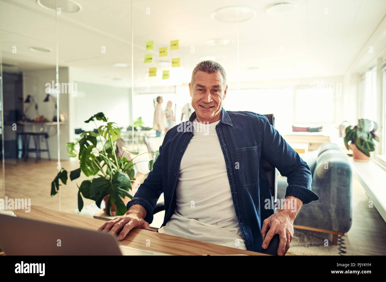 Reifen Geschäftsmann selbstbewusst lächelnd, während an seinem Schreibtisch sitzen in einem modernen Büro an einem Notebook arbeitet Stockfoto