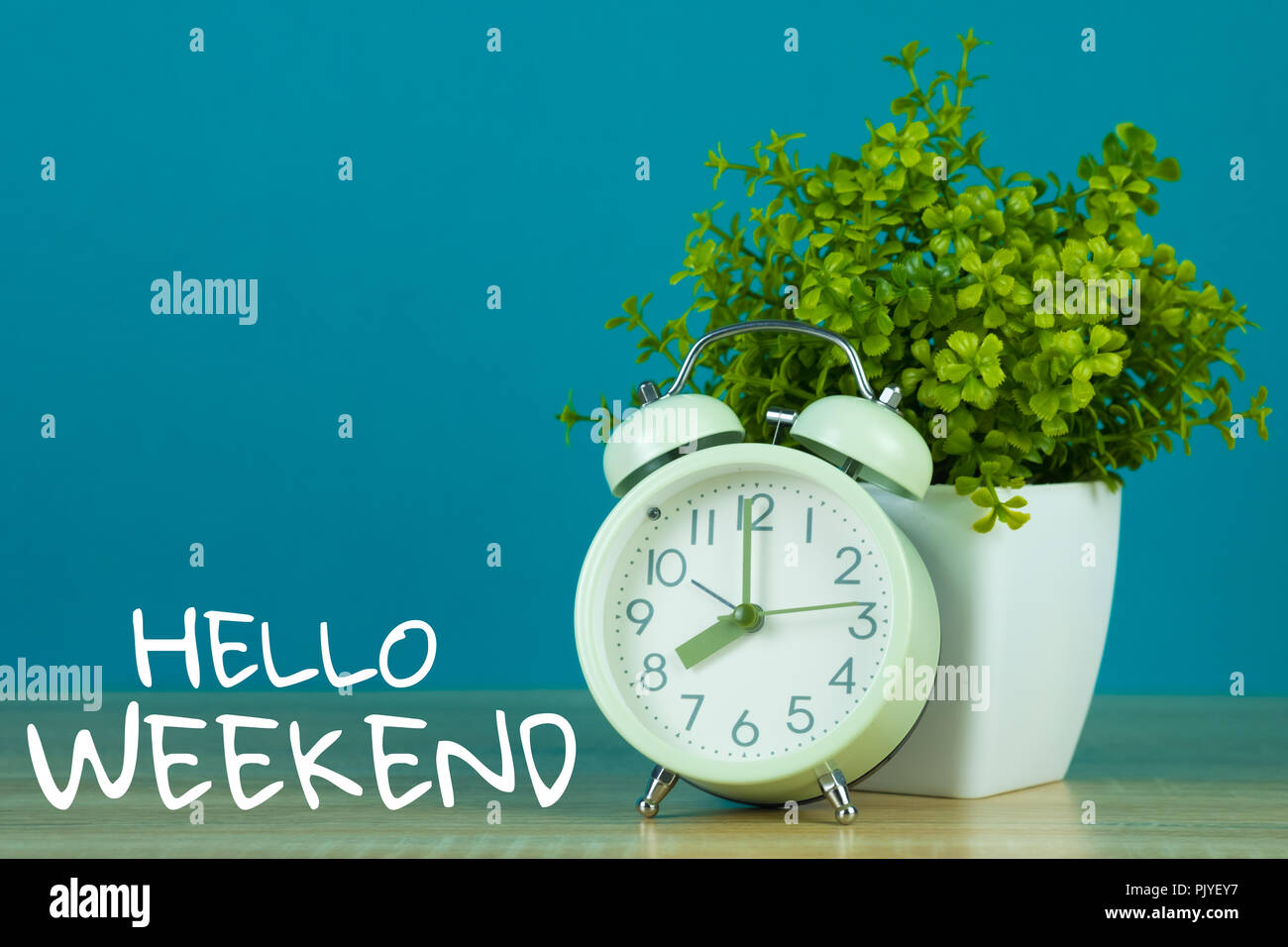 Hallo Wochenende Text auf grünem Hintergrund und kleinen Baum in weiße Vase mit Vintage Wecker auf Holztisch. Stockfoto