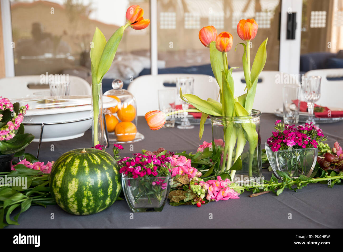 Ein Mittagstisch mit floralen Anzeige der Schönen orangen Blüten mit bunten rosa bis rötlich Arrangements im Vordergrund mit einem runden Wassermelonen & Trauben Stockfoto