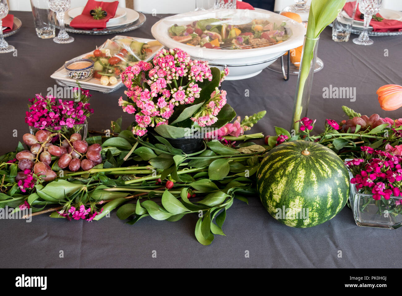 Ein Mittagstisch mit floralen Anzeige der Schönen orangen Blüten mit bunten rosa bis rötlich Arrangements im Vordergrund mit einem runden Wassermelonen & Trauben Stockfoto