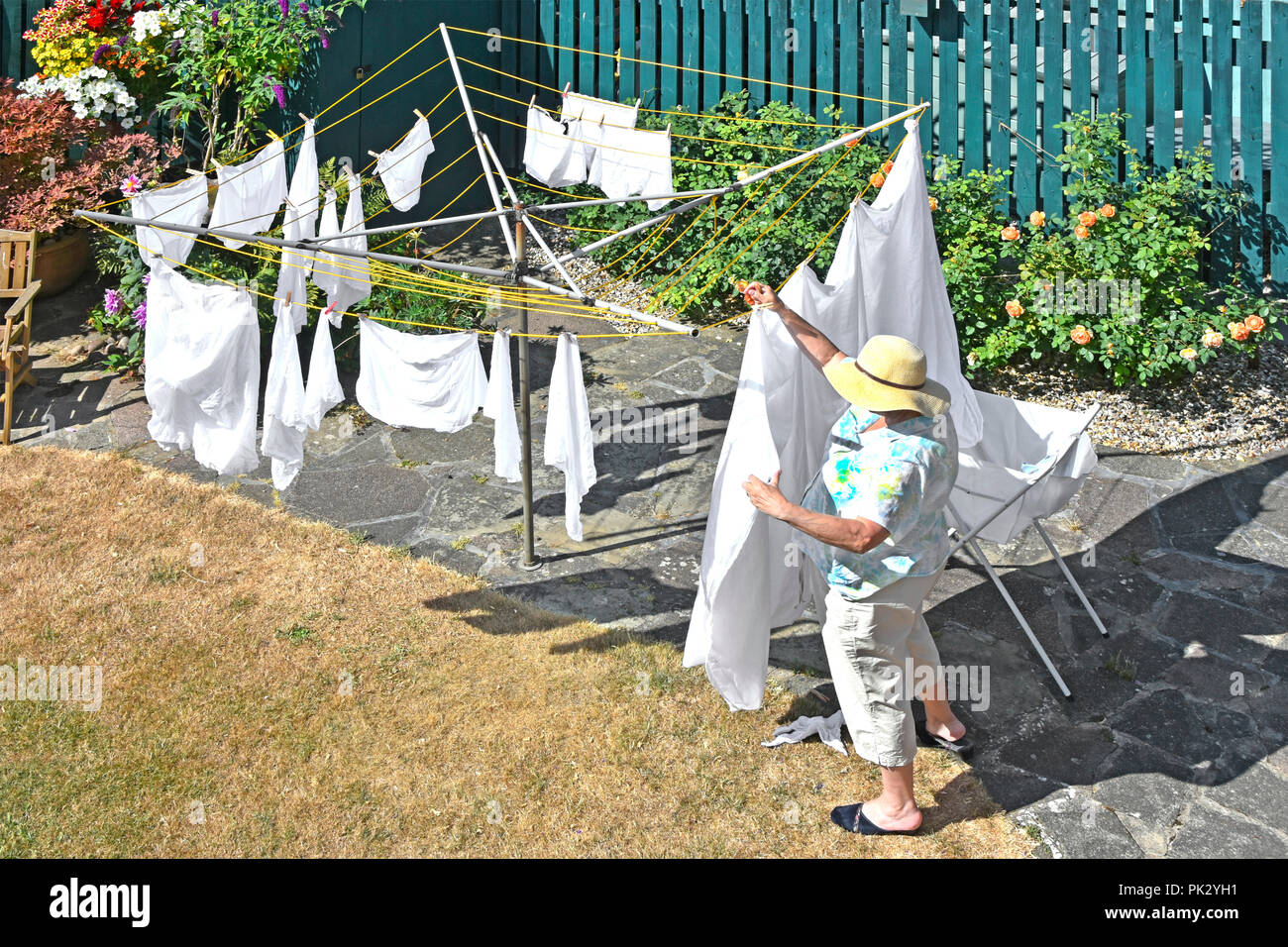 Hot 2018 Sommer auf geröstete trocken zurück Garten Rasen & reife ältere Frau mit Sonnenhut hängenden Waschen bei Rotary Wäscheleine England Großbritannien Stockfoto