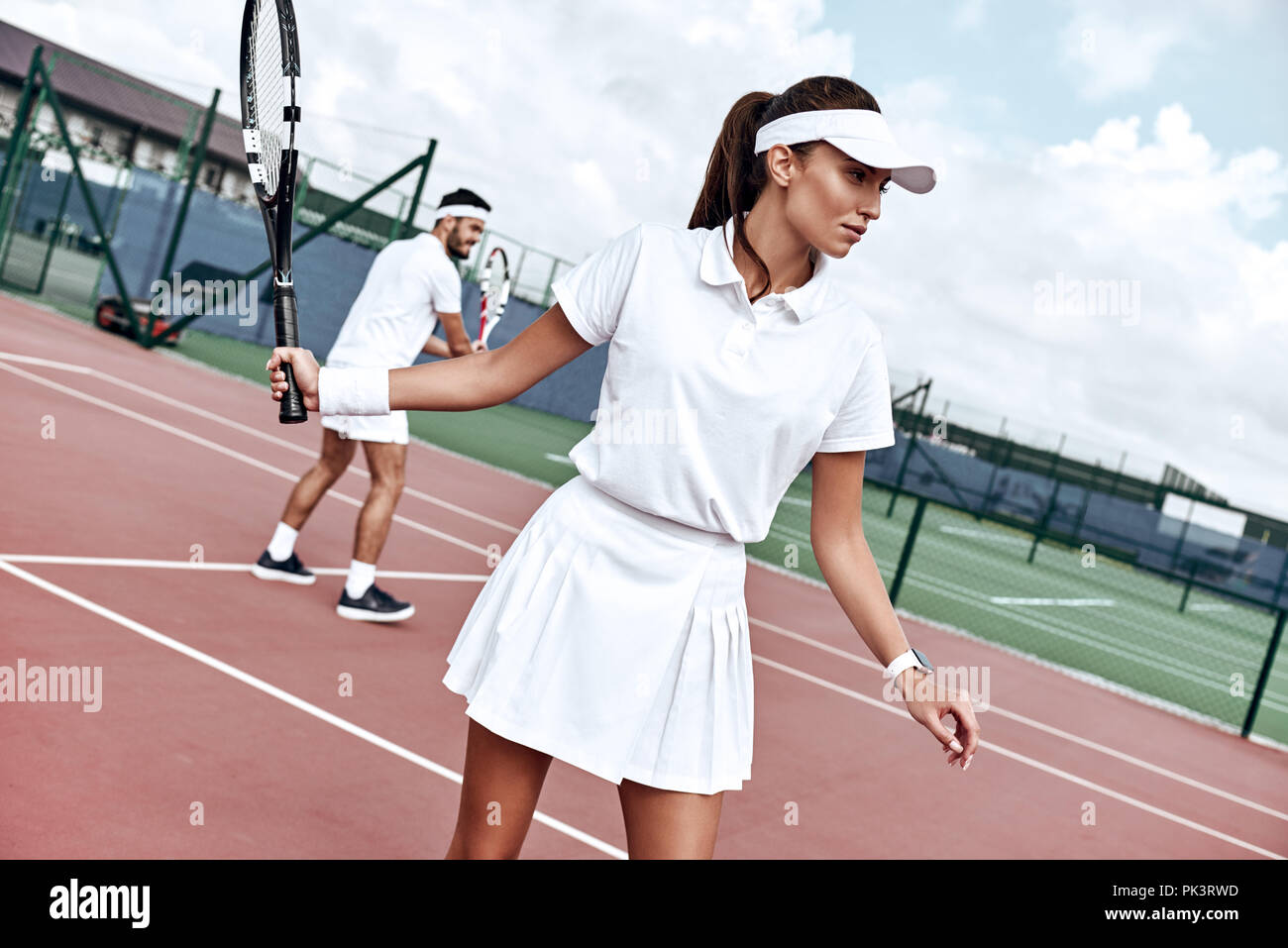 Sie spielen wie eine Mannschaft. Schöne junge Frau mit Tennisschläger und weg schauen beim Stehen auf einem Tennisplatz und mit Mann im Hintergrund Stockfoto
