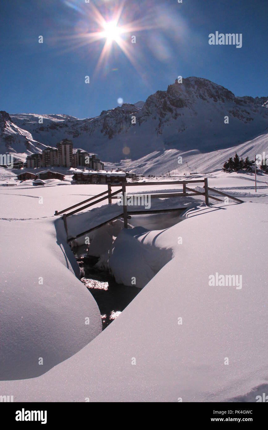 Sonnigen Wintertag, Le Lac, Tignes, Frankreich. Eine Brücke mit Schnee bedeckt, corssing einen kleinen zugefrorenen Fluss. Keine Anzeichen von schmelzendem Schnee unter der hellen Sonne Stockfoto