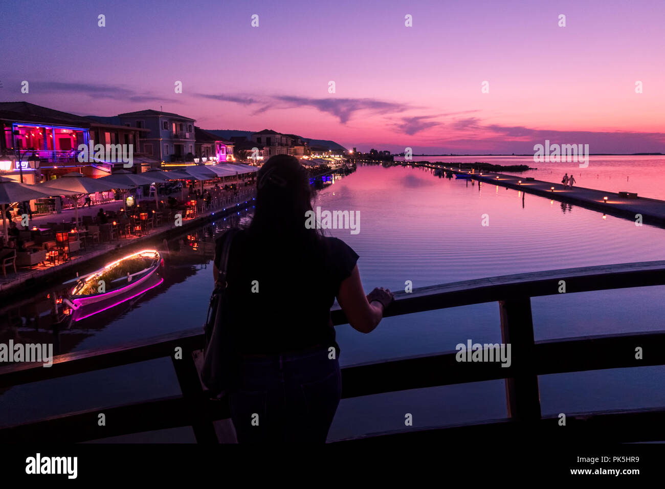 Frau mit dem Rücken zur Kamera, die den Sonnenuntergang auf der Marina-Brücke in Lefkada (Lefkas), Griechenland, beobachtet Stockfoto