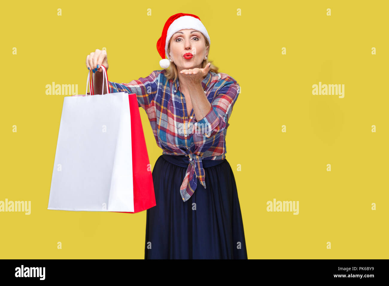 Portrait von bitte schön erwachsenen Frau in rot santa Kappe und kariertem Hemd, die hielten Einkaufstaschen und senden Sie küssen, am Cam suchen Stockfoto
