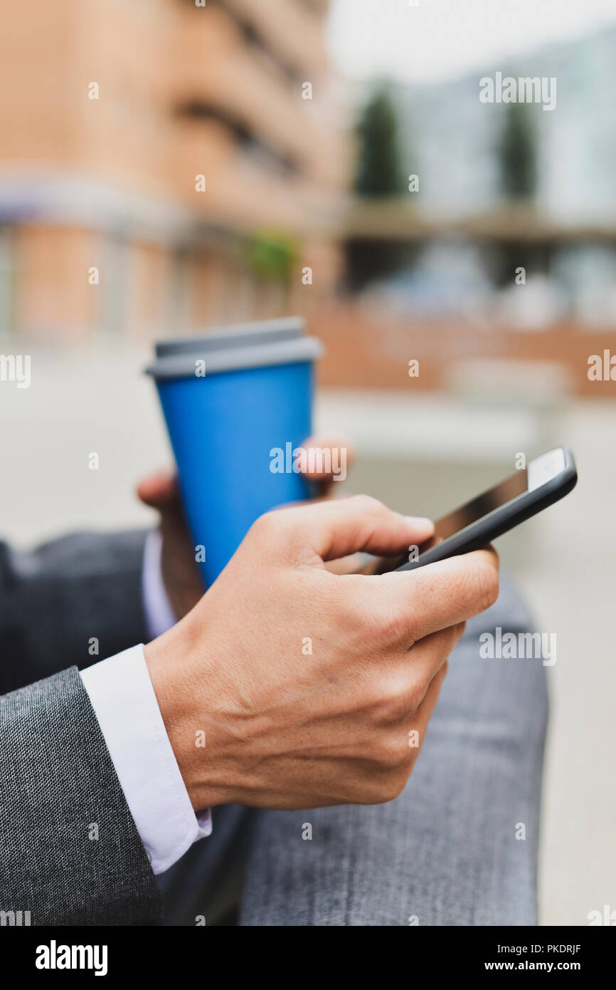 Nahaufnahme eines jungen kaukasischen Geschäftsmann in einem eleganten Grau mit einem Smartphone Anzug und einen Kaffee im Freien Stockfoto