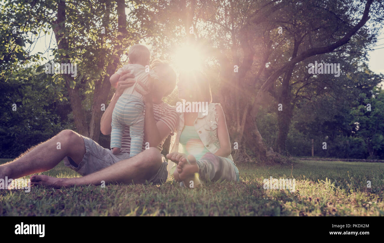 Schöne Familie Moment - Junges Paar sitzt im Gras in einem Park mit Bäumen während Vater ihr Kleinkind Kind hält durch ein helles Sonnenlicht beleuchtet. Mit ret Stockfoto