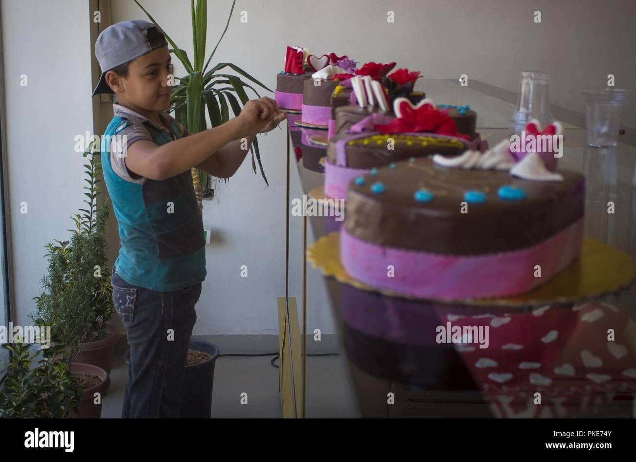 Ein Junge verleiht eine Hand durch die dekorierten Kuchen aus der Bäckerei auf der Sweet Shop in Manbij, Syrien, 26. Juli 2018. Geschäfte, Restaurants und Bäckereien sind in Manbij wie Einheimische bereiten zweiten Jahrestag der Stadt der Befreiung von ISIS durch die SDF zu feiern. Stockfoto