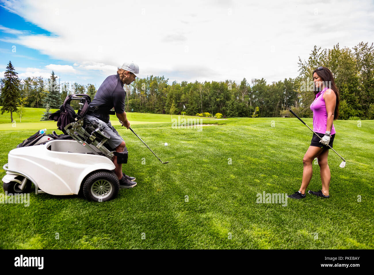 Ein körperlich behinderter Golfer, mit einem spezialisierten Rollstuhl, trifft den Golfball mit seinem Golfschläger auf dem Golf-Grün, wie eine Golfspielerin steht ... Stockfoto