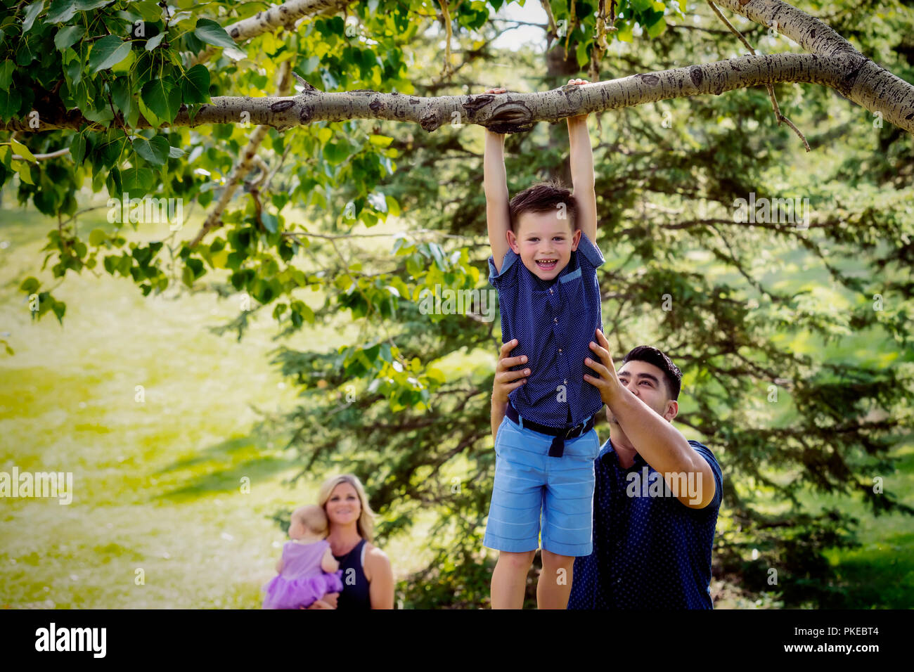 Eine Mutter und Baby-Uhr, während der Vater hält seinen Sohn hoch, um ihm zu helfen, hängen an einem Baum Ast während eines Familie... Stockfoto