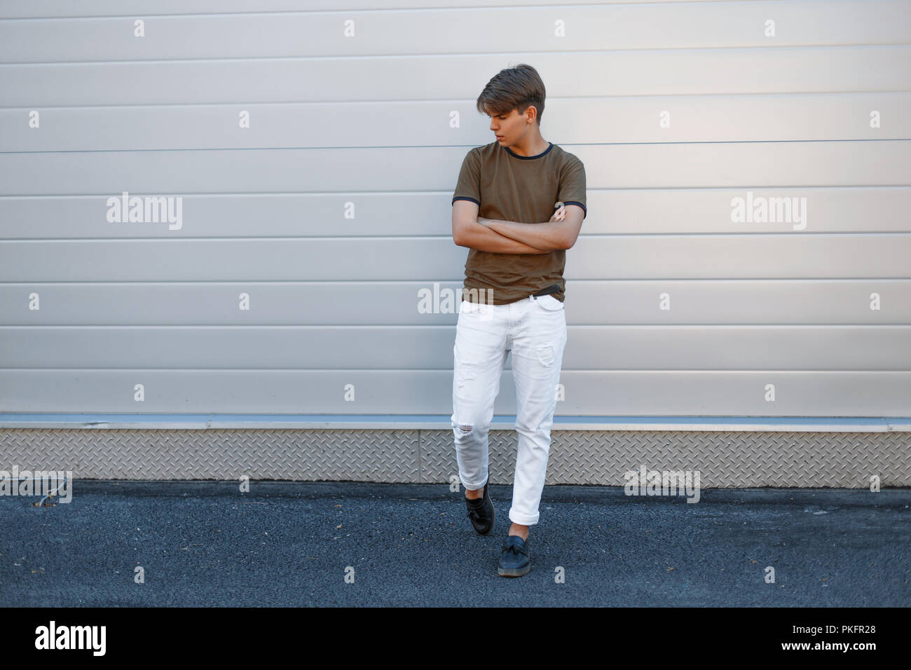 Schönes Modell von einem Mann im vollen Wachstum in einem Fashion T-Shirt mit weißen Hosen und schwarze Schuhe in der Nähe der Graue glänzende Wand Stockfoto