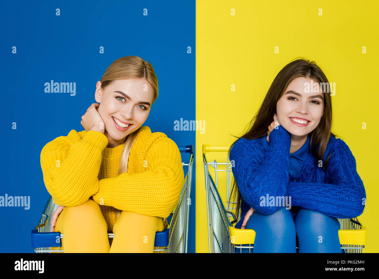 Attraktive junge Mädchen Spaß in Shopping Carts auf blauen und gelben Hintergrund sitzen Stockfoto