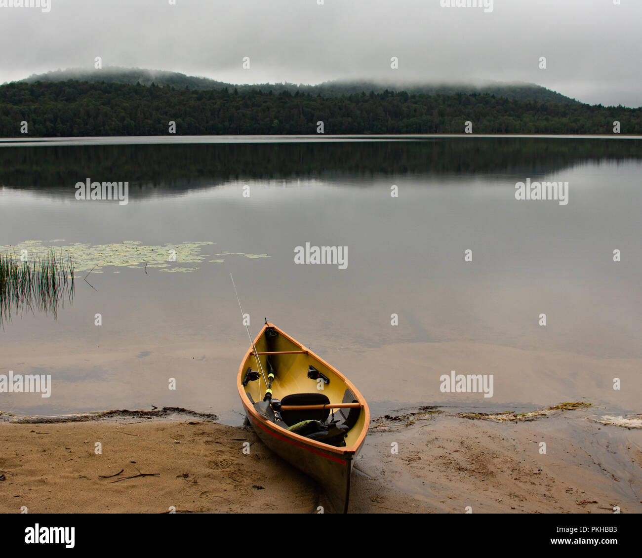 Ein leichtes Kanu mit Fly Fishing Rod. Paddel und Schwimmweste an einem Sandstrand auf einem Adirondack Mountains, NY Wüste See auf einem nebligen Morgen Stockfoto