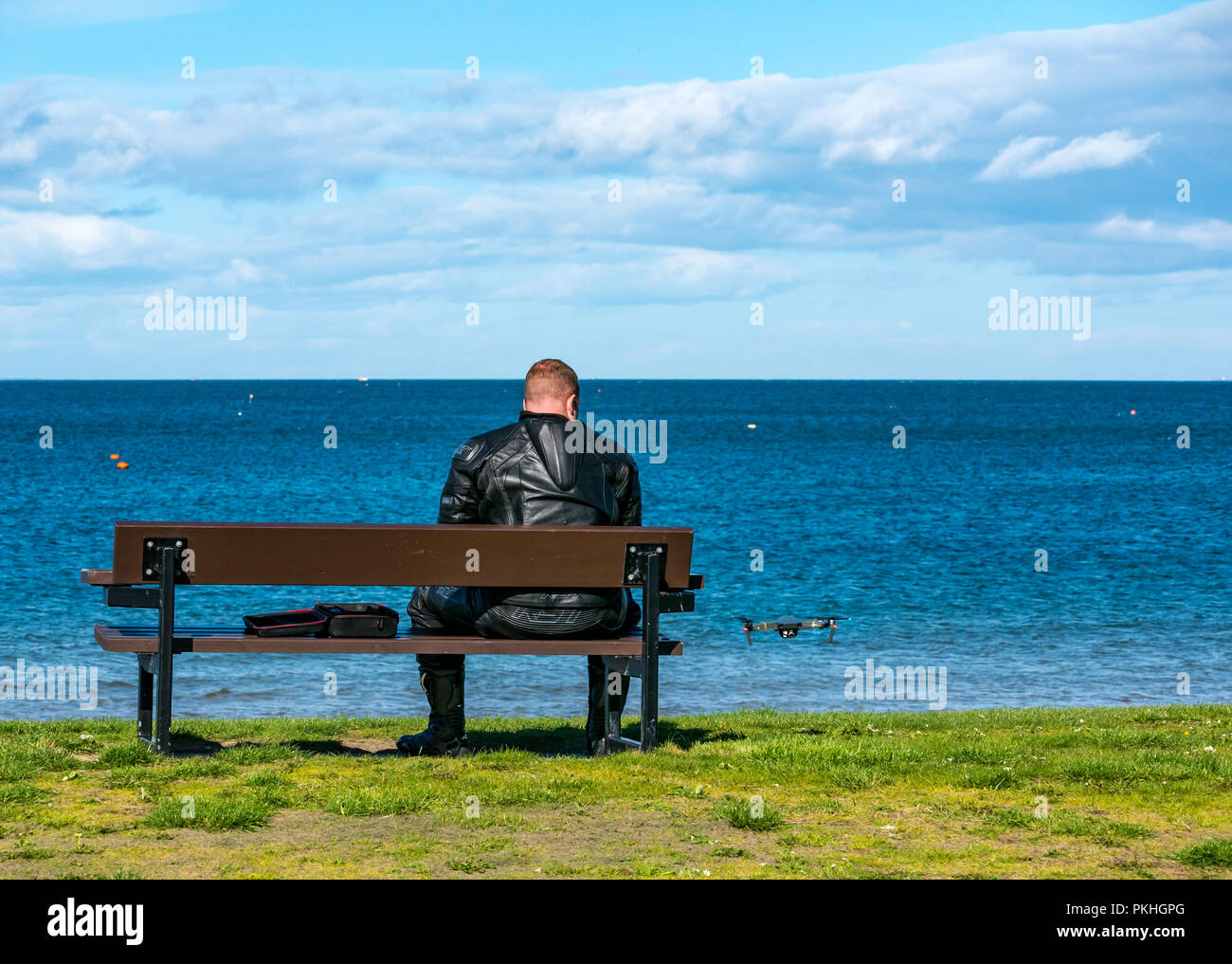 Mann in Motorrad Leder sitzt auf der Bank flying Drone, North Berwick, East Lothian, Schottland, Großbritannien Stockfoto