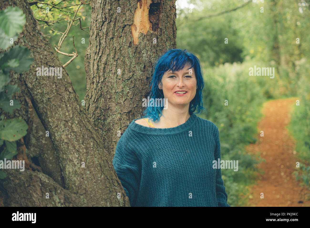 Eine blaue behaarte Frau, trägt einen blauen Pullover, lehnt sich gegen einen Baum auf einem Waldweg Stockfoto