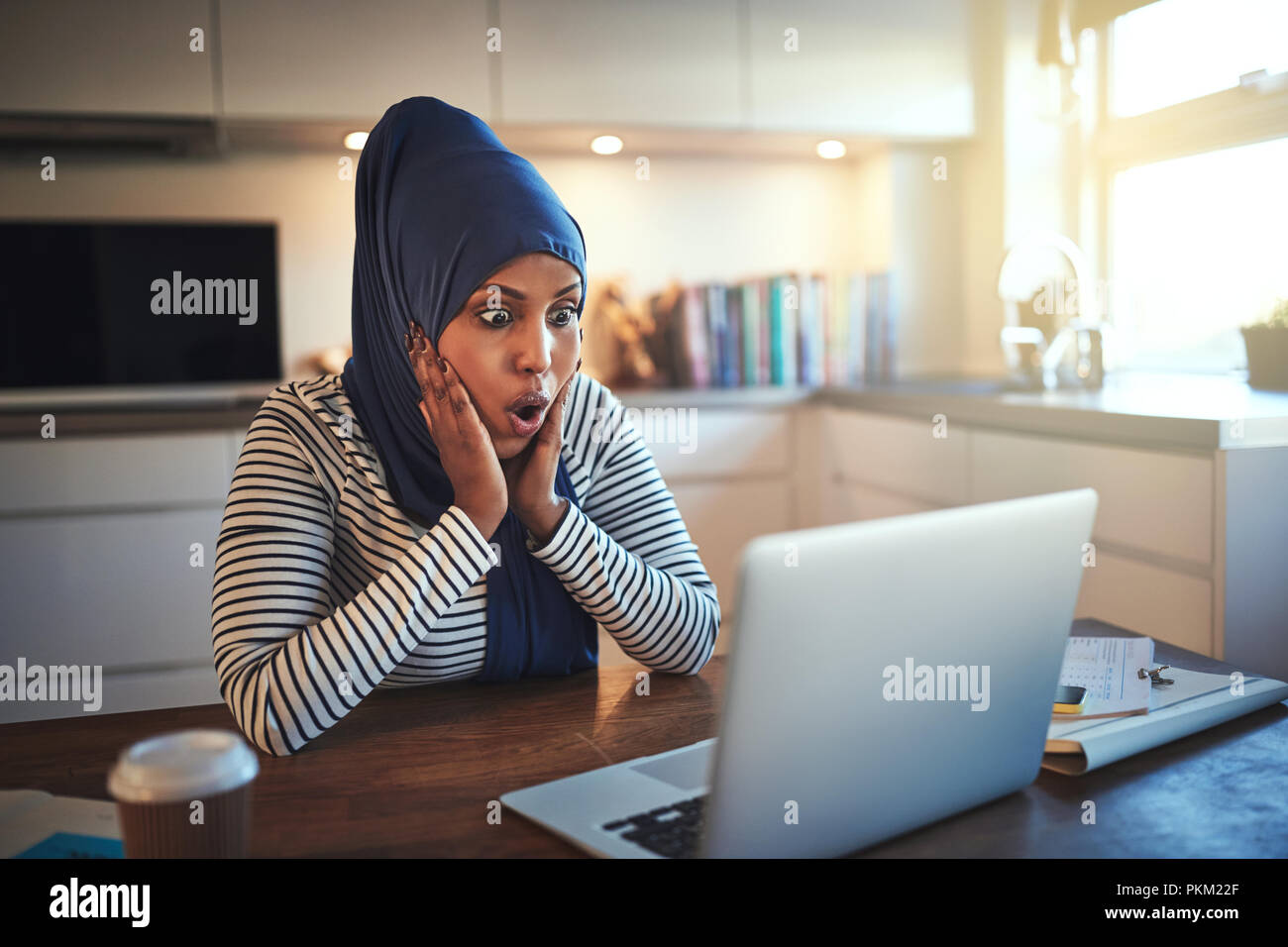 Junge arabische weibliche Unternehmer Tragen eines hijab überrascht, während an ihrem Küchentisch, der an einem Notebook arbeitet Stockfoto