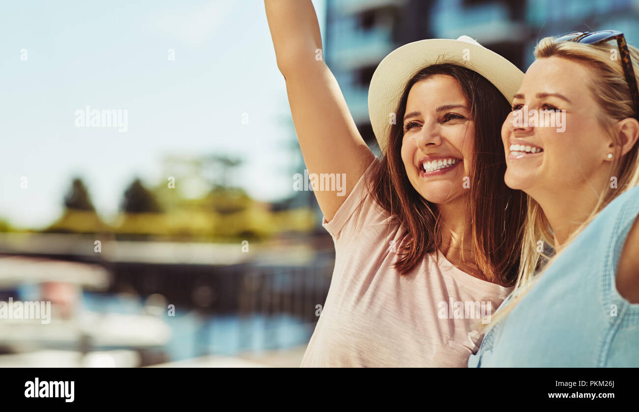Zwei lachende Junge weibliche Freunde stehen Arm in Arm zusammen mit einem sonnigen Sommertag in der Stadt Stockfoto