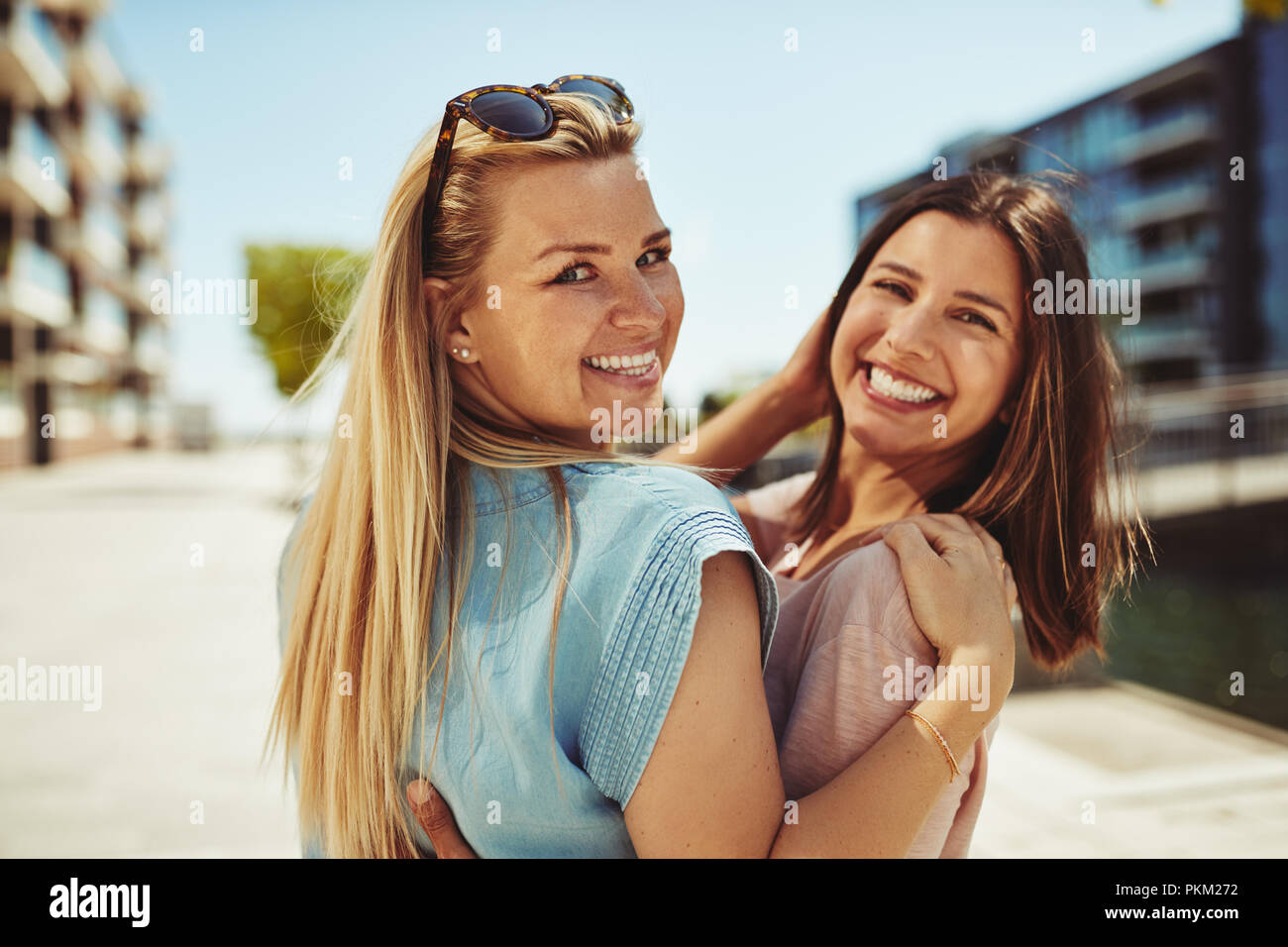 Zwei lächelnde junge weibliche Freunde über die Schulter schauen beim Gehen Arm in Arm zusammen in der Stadt im Sommer Stockfoto