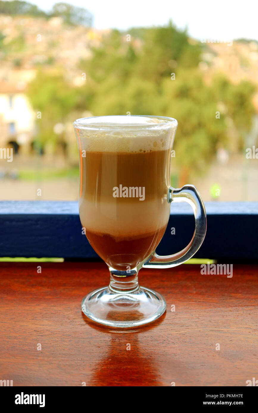 Vertikale Foto von heißen Cappuccino in einem transparenten Schale an der Terrasse mit unscharfen alten Stadtplatz von Cusco, Peru im Hintergrund serviert. Stockfoto